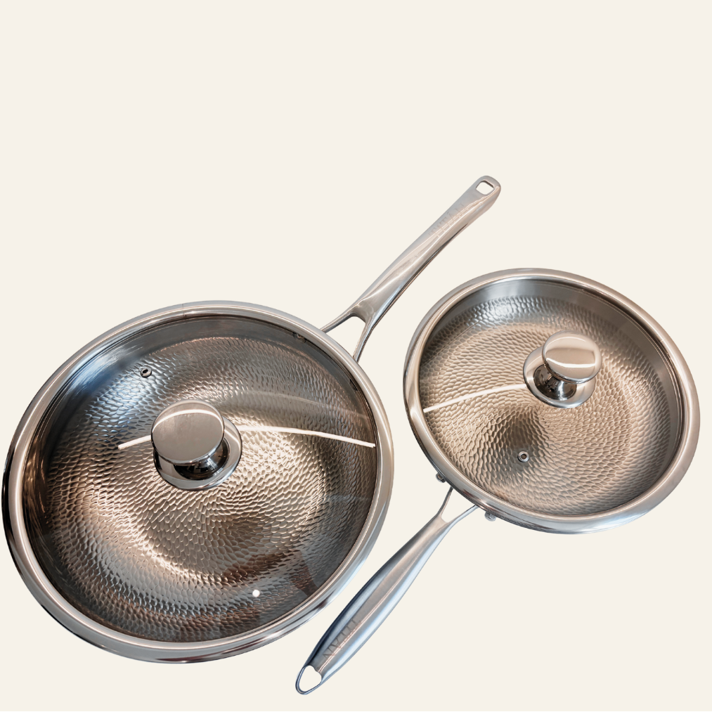 Two titanium saucepans with matching glass lids and metal handles, displayed on a solid off-white background.
