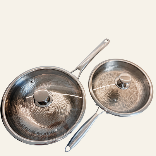 Two titanium saucepans with matching glass lids and metal handles, displayed on a solid off-white background.