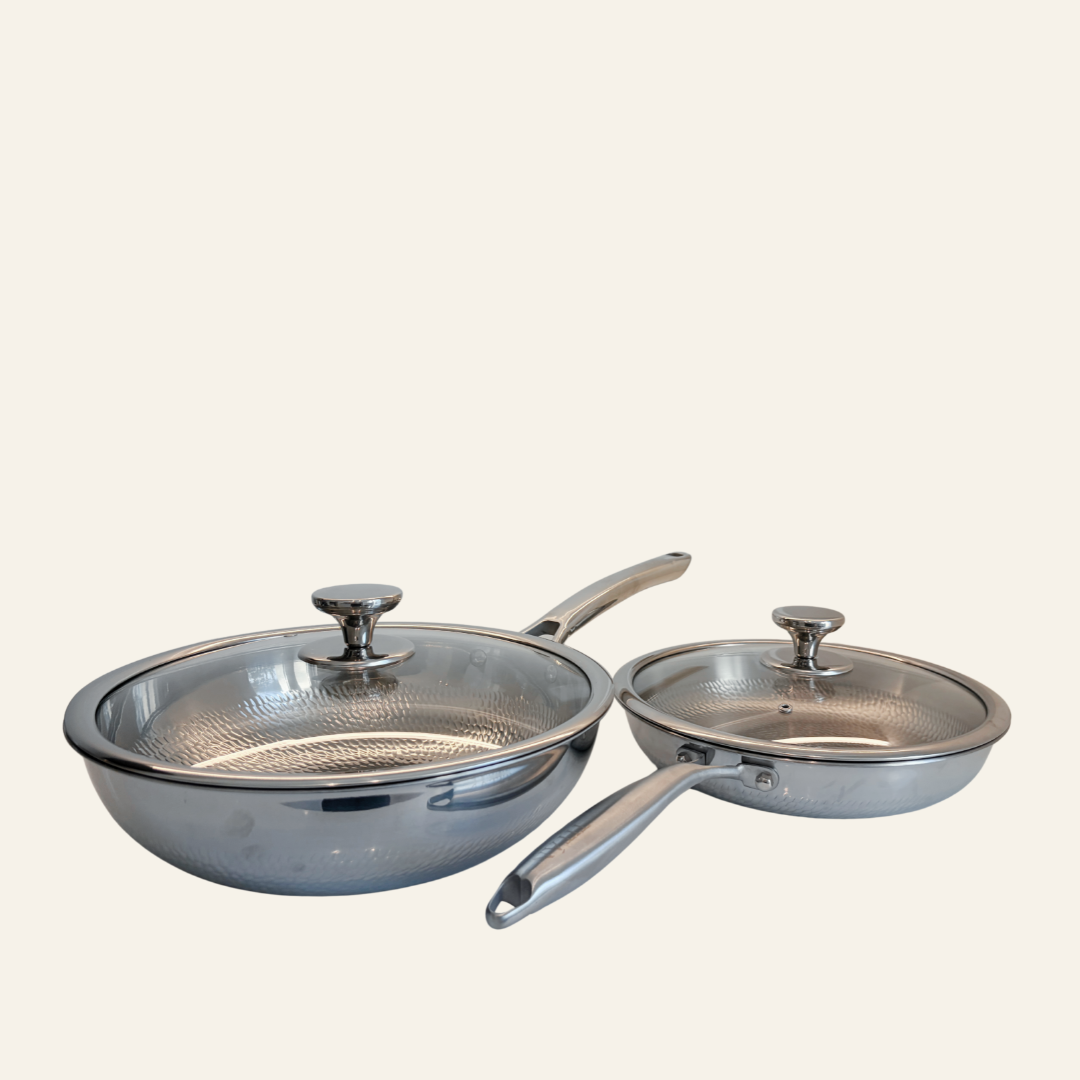 Two titanium cookware pans with glass lids, one larger deep pan and one smaller skillet, shown on a plain light beige background.